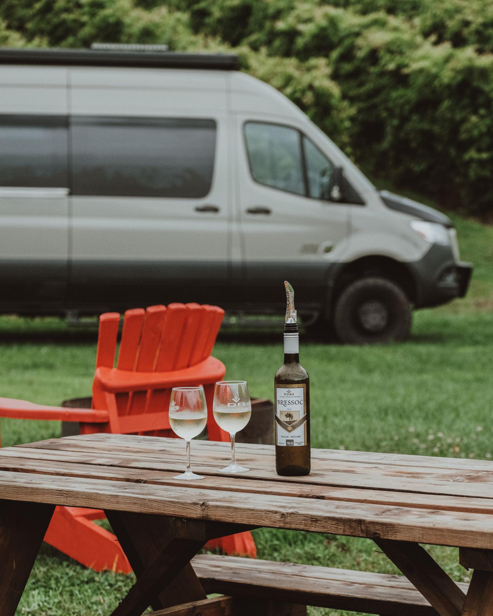 image1 Bouteille de vin blanc avec deux coupes sur une table de pique-nique, avec une van en arrière-plan.