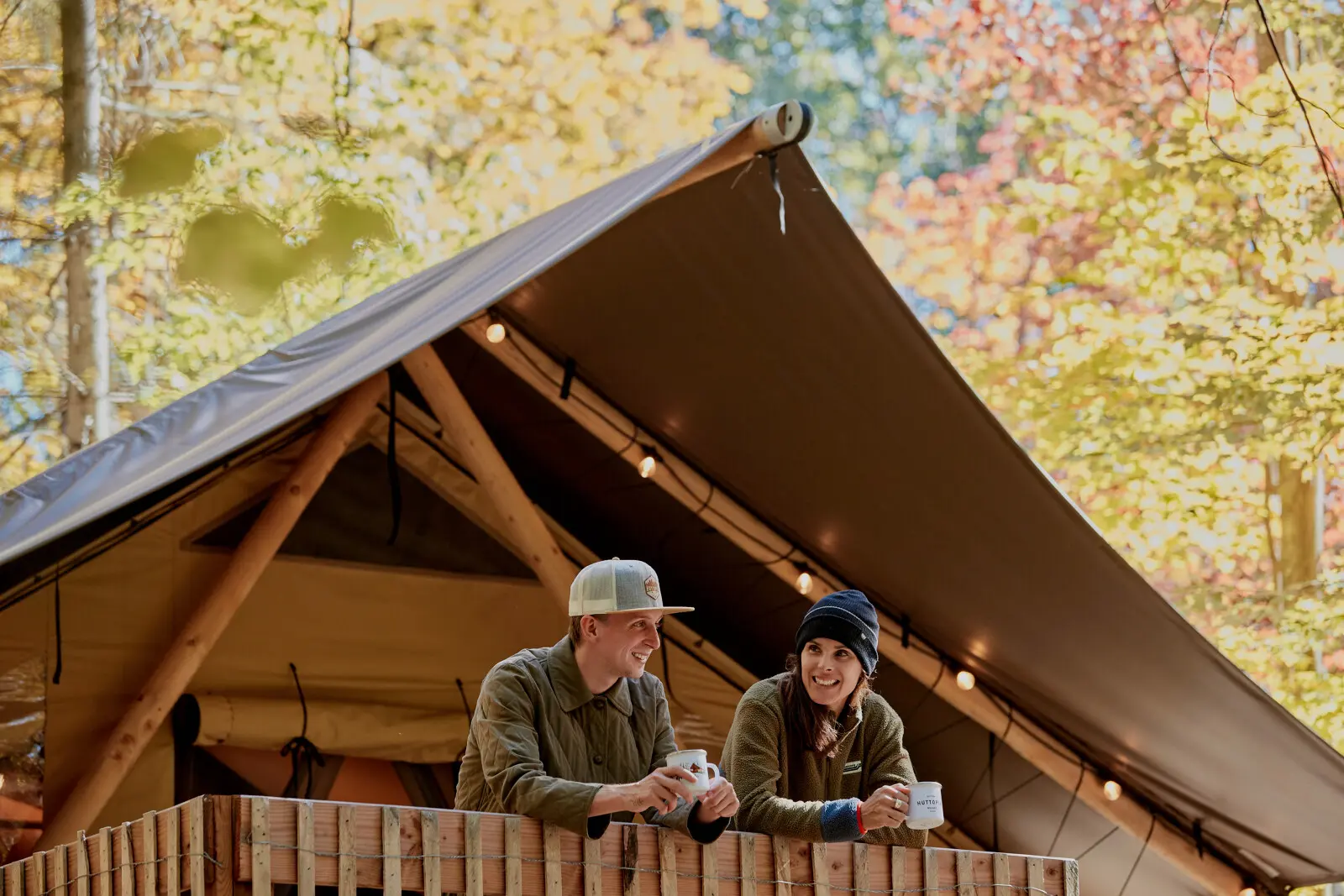 experience glamping route des vins brome missisquoi Couple souriant sur un balcon de glamping, tasses à la main, avec forêt automnale. Dans un hébergement d'Huttopia Sutton.