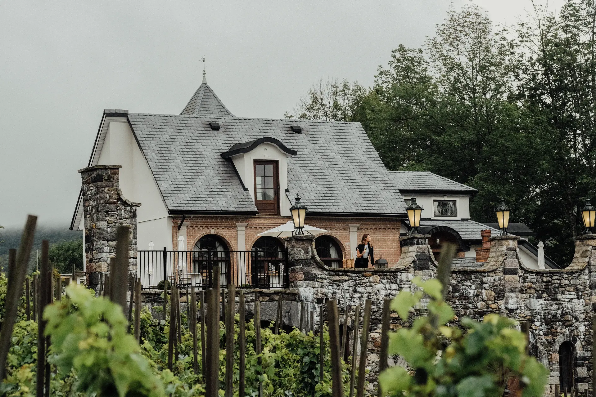 vignoble route des vins brome missisquoi 1 Bâtiment du vignoble Château Ste-Agnès, en pierre et ardoise, femme sur balcon avec verre de vin, surplombant les vignes de la Route des vins Brome-Missisquoi.